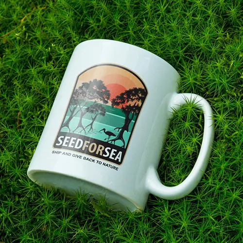 Mug blanc avec logo Seed for Sea représentant des mangroves, la mer et un coucher de soleil, posé sur un tapis de mousse verte.